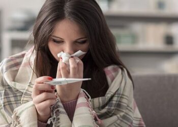 Sick young woman at home on the sofa, she is covering with a blanket, taking temperature and blowing her nose with a tissue