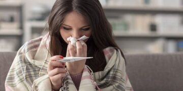 Sick young woman at home on the sofa, she is covering with a blanket, taking temperature and blowing her nose with a tissue